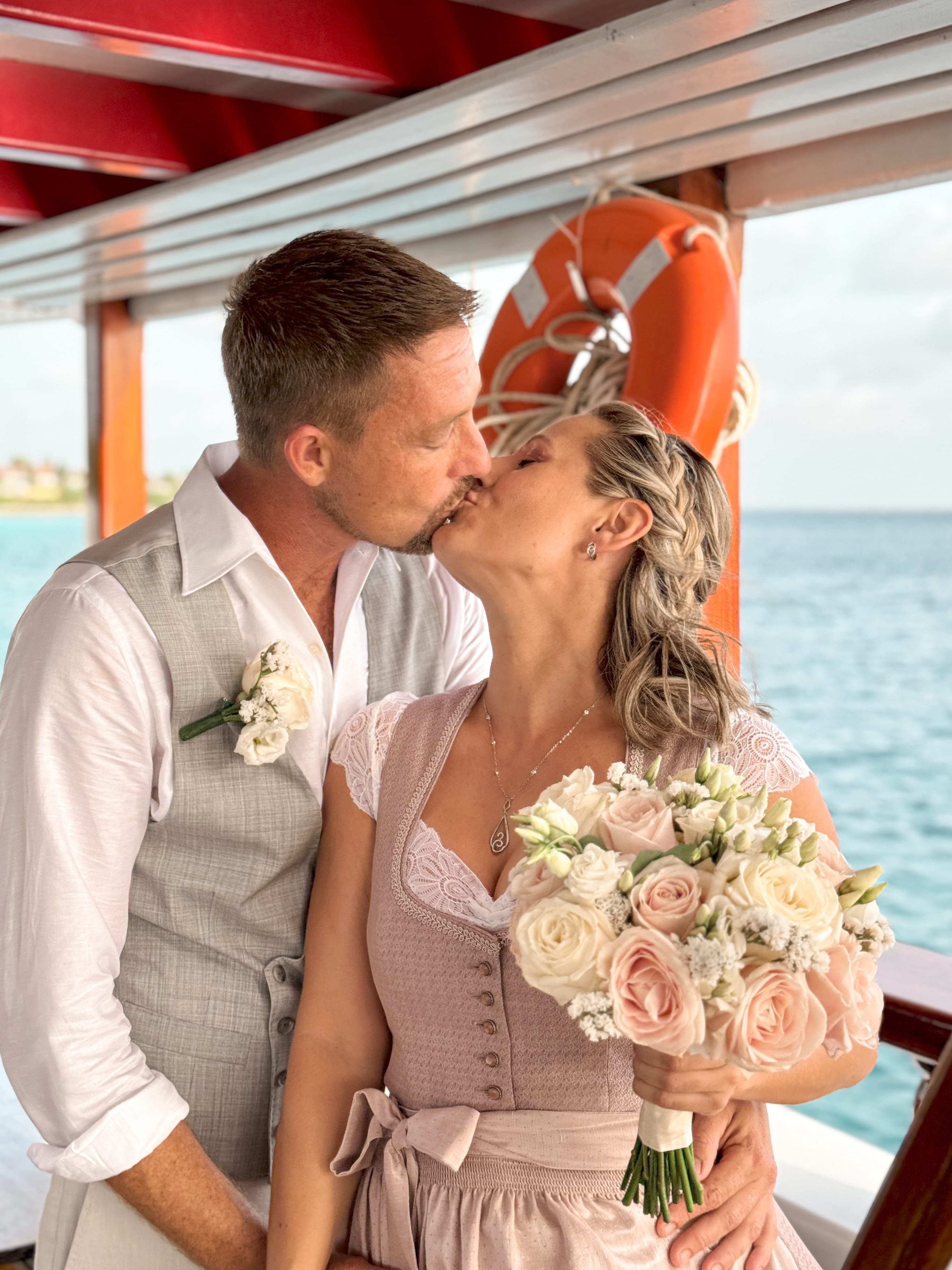 A newly married couple kissing on deck, the bride holding a bouquet of roses
